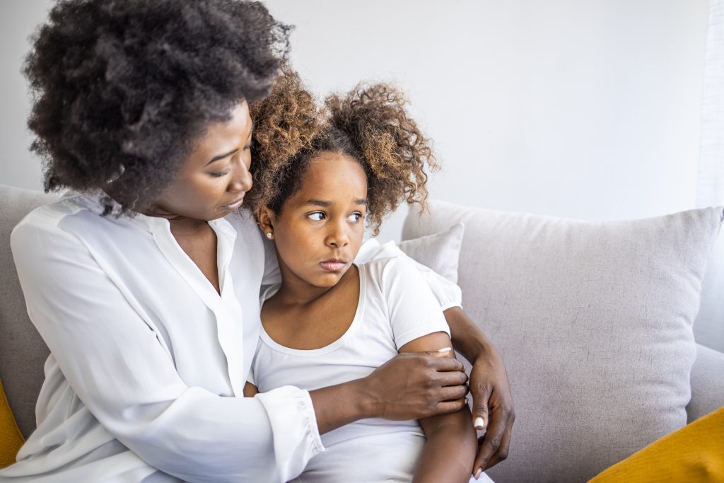 A mother comforts her upset child while they sit on a sofa.